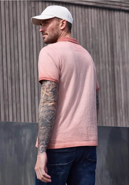 Ein Mann steht seitlich vom Betrachter, trägt rosa Polo-Shirt, Jeans, weiße Kappe und tätowierte Arme.