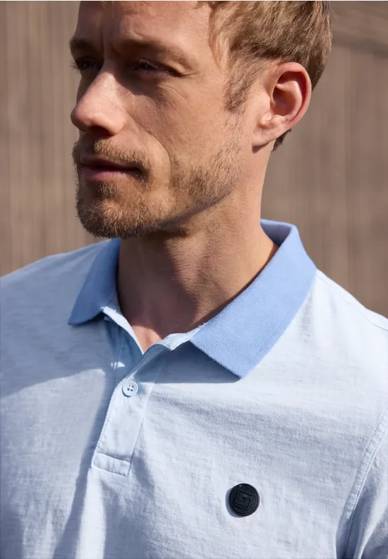 Nahaufnahme eines Mannes mit Kurzhaarschnitt und hellblauem Poloshirt vor Holzwand.