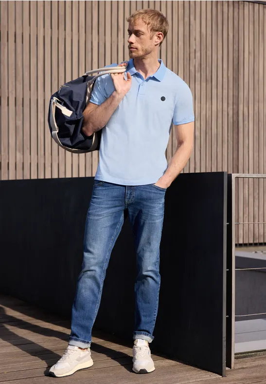Ein Mann mit Poloshirt, Jeans und Tasche steht vor moderner Architektur.