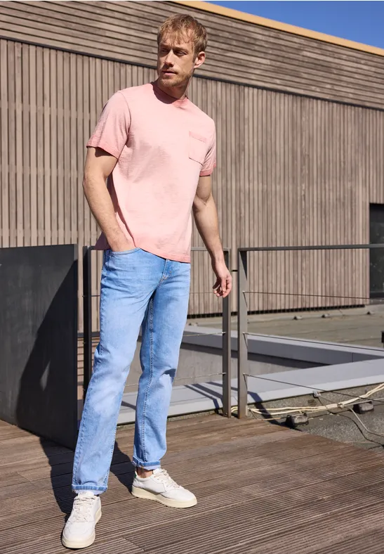 Een man met roze T-shirt, lichtblauwe jeans en witte sneakers staat op een houten terras bij een gebouw.