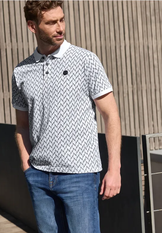 Ein junger Mann trägt ein gemustertes weißes Poloshirt und blaue Jeans vor einer Holzfassade.
