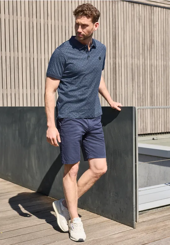 Ein Mann lehnt auf einer Dachterrasse an einem Geländer, trägt blaues Polo, Shorts und weiße Sneaker.