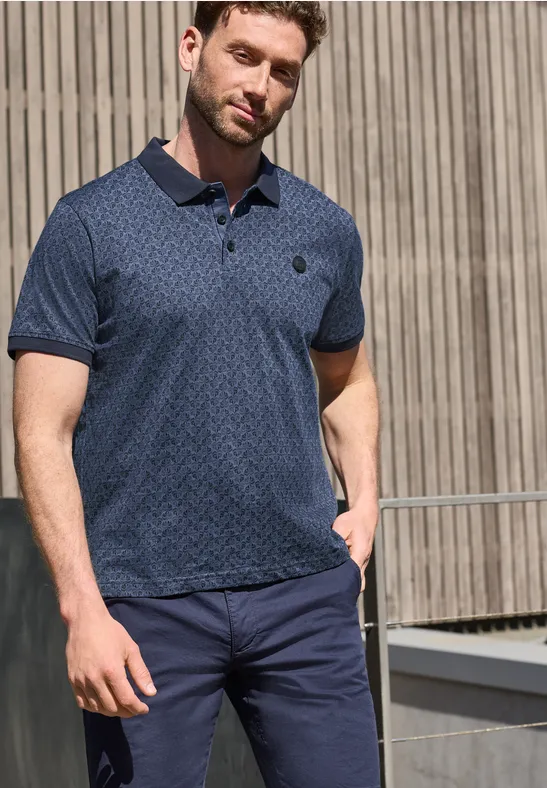 Ein Mann steht vor einer Holzfassade und trägt ein gemustertes blaues Poloshirt und dunkelblaue Hose.