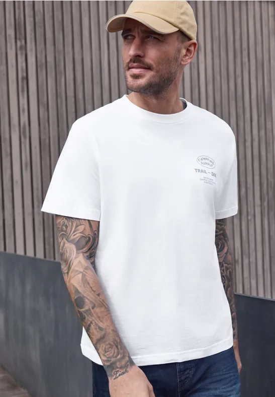 Ein Mann in weißem T-Shirt mit auffälligen Arm-Tattoos steht vor einer Holzlattenwand.