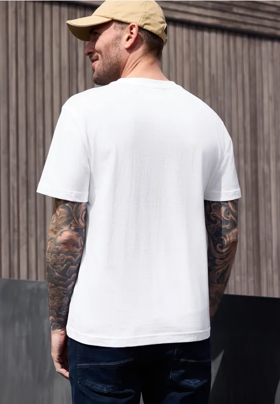 Mann mit Baseballcap, weißem T-Shirt, dunklen Jeans und tätowierten Armen von hinten vor Holzwand.