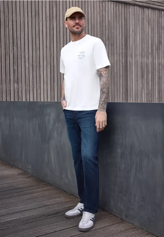 Ein tätowierter Mann steht in weißem T-Shirt, blauen Jeans und weißen Sneakern vor einer Holz- und Stahlwand.