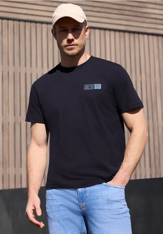 Ein Mann in schwarzem T-Shirt und blauer Jeans steht vor einer Holzwand.