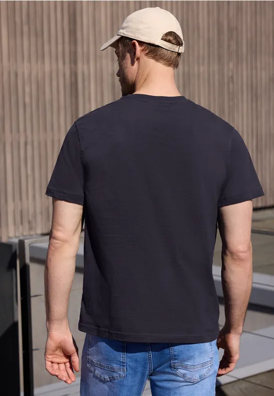 Ein Mann steht mit dem Rücken zur Kamera, trägt dunkelblaues T-Shirt, blaue Jeans und helle Kappe.