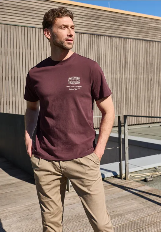 Ein junger Mann trägt ein T-Shirt und Chinos und steht vor einer Holzfassade auf einer Dachterrasse.