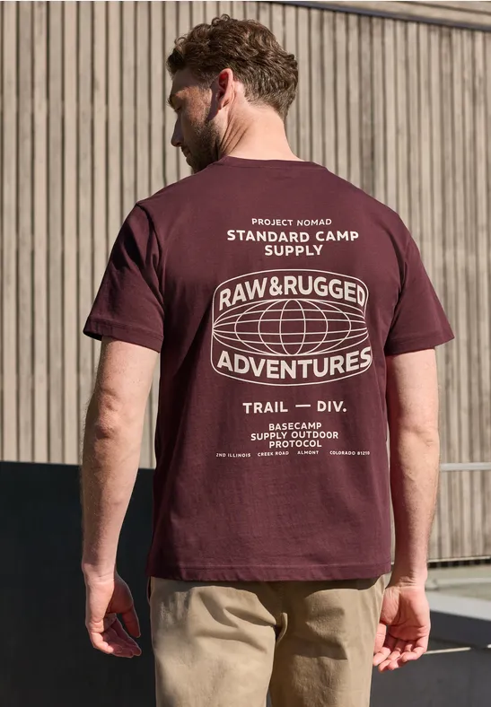 Ein Mann in beiger Hose vor Holzfassade trägt von hinten ein bordeauxfarbenes T-Shirt mit weißem 'Raw & Rugged Adventures'-Aufdruck.