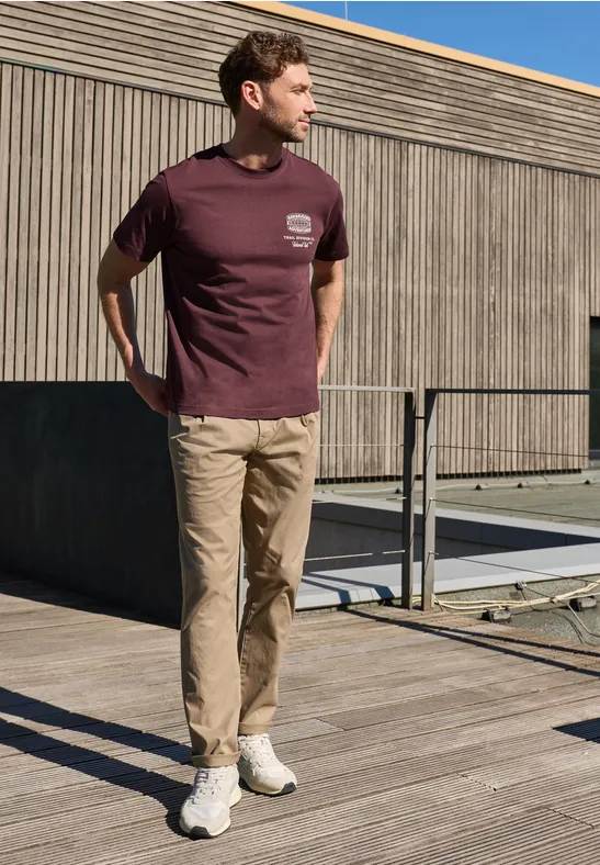 Ein Mann steht auf einer Holzterrasse, trägt ein bordeauxfarbenes T-Shirt, eine beige Hose und weiße Sneaker.
