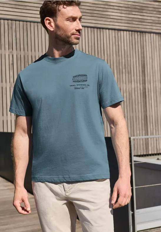 Ein Mann in blau-grünem T-Shirt und heller Hose steht vor einer modernen Holzfassade.