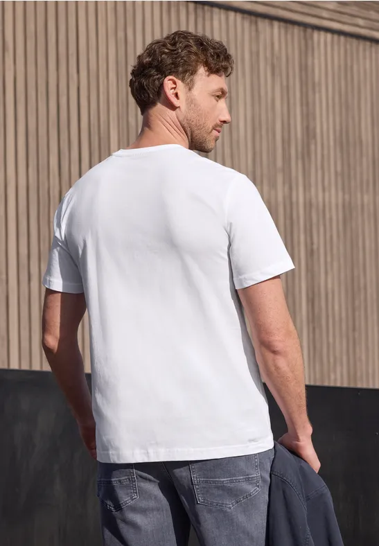Ein Mann steht mit weißem T-Shirt und Jeans vor einer Holzwand und hält eine Jacke in der Hand.