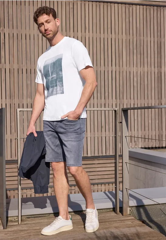 Ein Mann in weißem T-Shirt und grauen Shorts steht auf einem Balkon und hält eine Jacke.