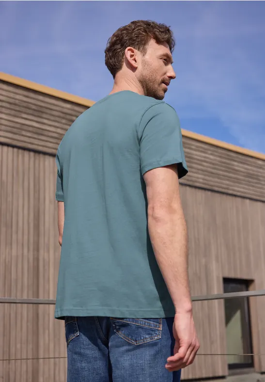 Ein Mann steht mit dem Rücken zur Kamera, trägt ein türkises T-Shirt und Jeans vor einer Holzfassade.
