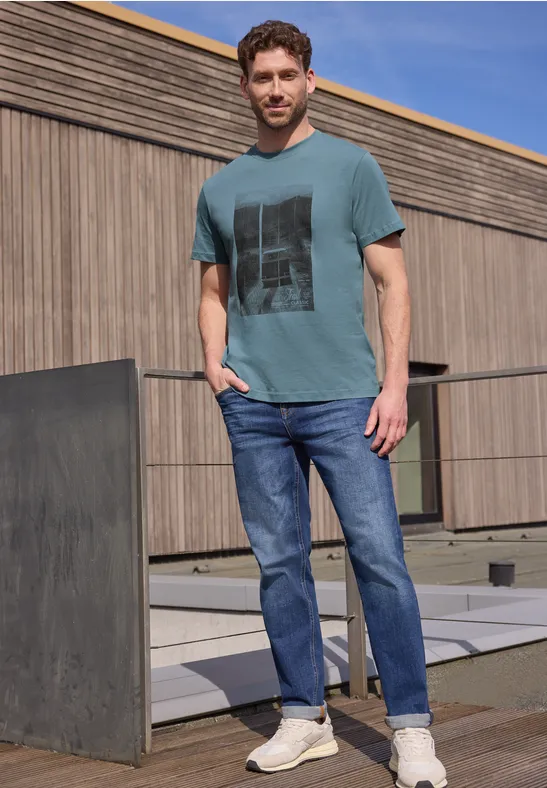 Ein Mann in grünem Print-T-Shirt, blauen Jeans und Sneakern steht auf einer Betonterrasse vor einer Holzfassade.