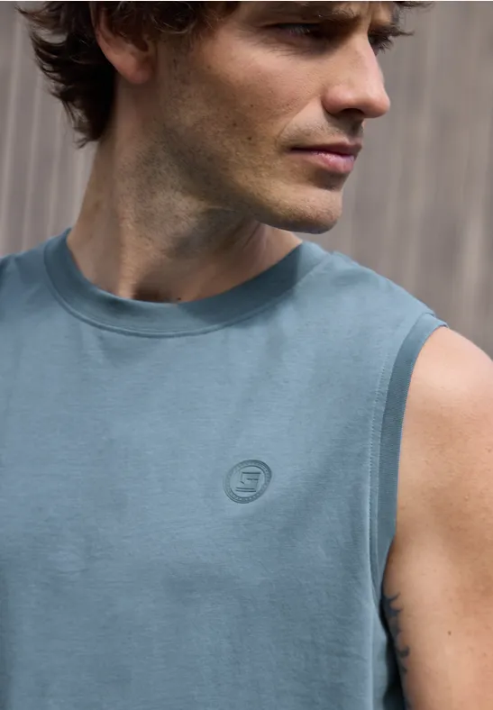 Oberkörper eines Mannes in ärmellosem blauen Shirt vor unscharfem Hintergrund.