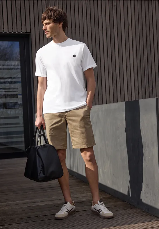 Een man in wit T-shirt, beige short en witte sneakers houdt een zwarte tas vast voor een moderne gevel.