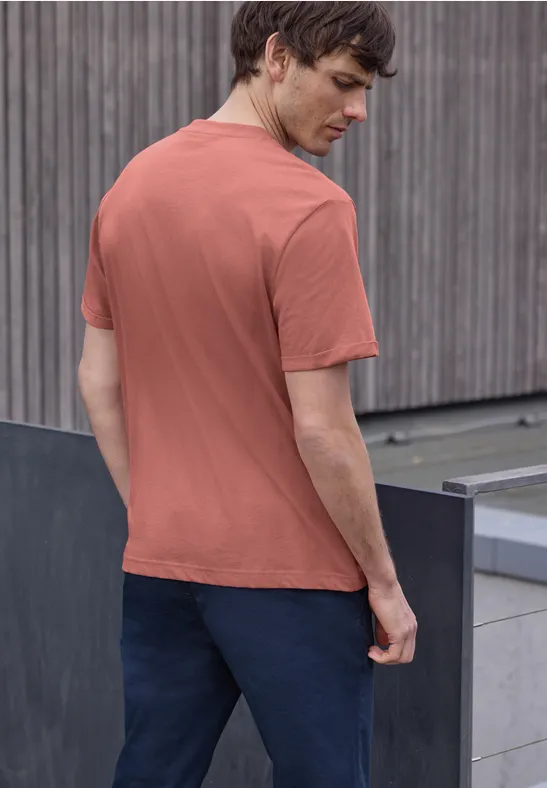 Een man van achteren in een baksteenrood t-shirt en donkere broek voor een houten gevel.