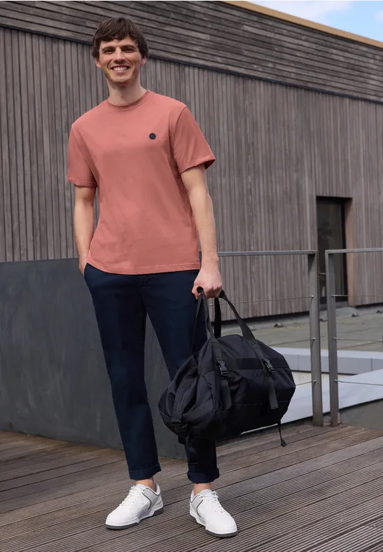 Een man in een koraal T-shirt en donkere broek draagt een zwarte reistas op een houten terras.