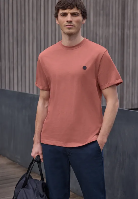 Een man draagt een roestkleurig t-shirt en donkere broek, met een zwarte tas voor een houten wand.