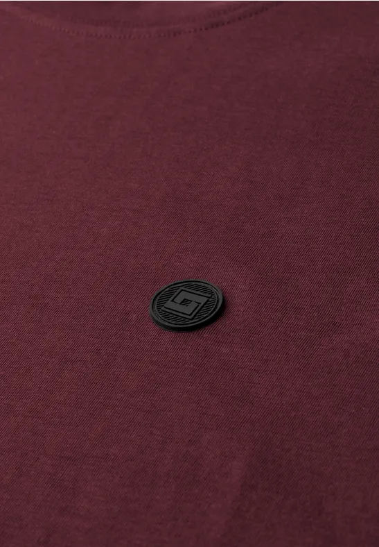 Een bordeauxrood T-shirt met een klein, zwart rond embleem op de borst.