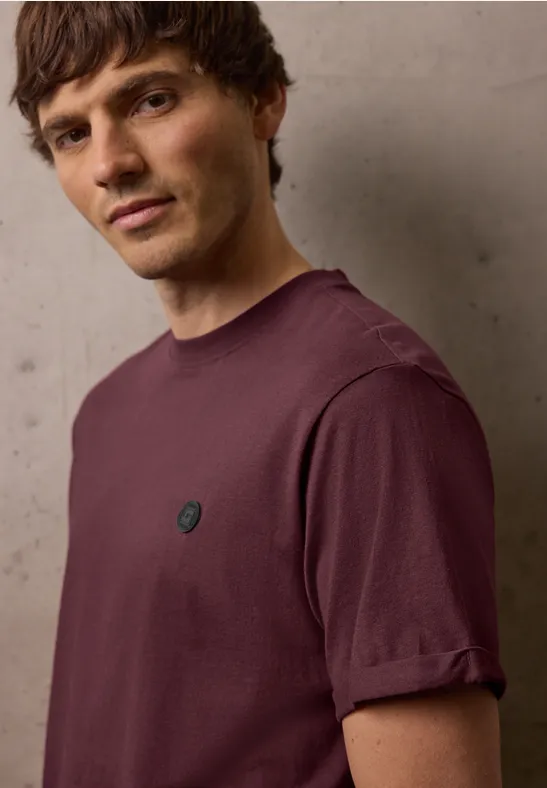 Een man draagt een eenvoudig paars T-shirt met opgerolde mouwen en een klein rond logo op de borst.