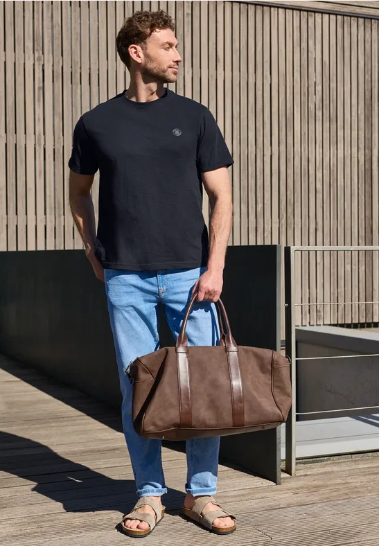 Ein Mann in schwarzem T-Shirt und Jeans hält eine braune Lederreisetasche vor einer Holzwand.