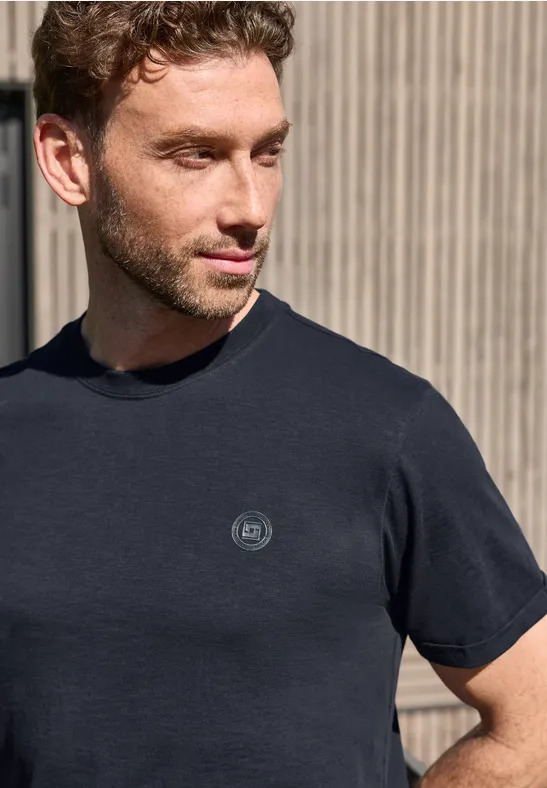 Ein Mann in schwarzem T-Shirt mit kleinem Logo steht vor einer schlichten, vertikal gestreiften Wand.