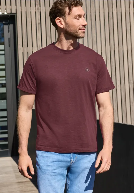 Ein Mann trägt ein weinrotes T-Shirt und Jeans, steht vor Holzpaneelen im Freien.