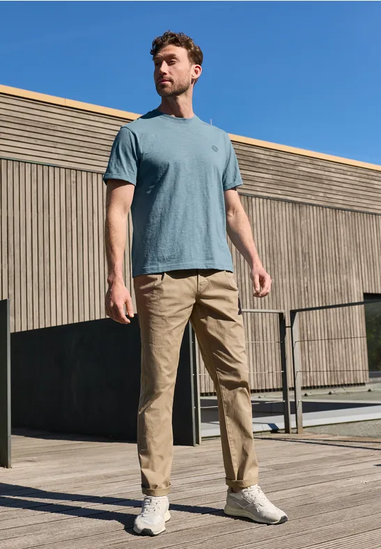 Mann mit blauem T-Shirt und beigen Hosen steht vor moderner Holzfassade.