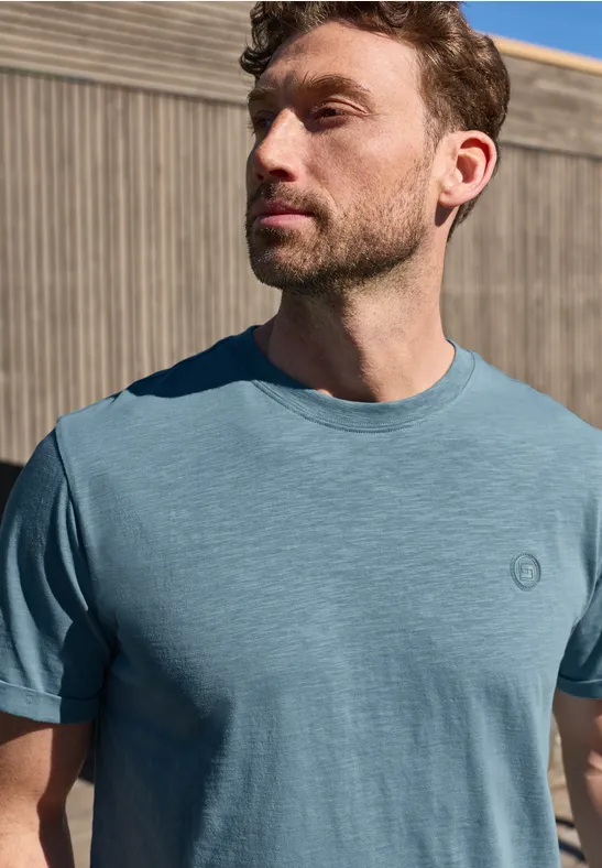 Mann steht vor Holzwand in blauem T-Shirt, schaut seitlich.