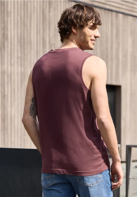 Ein Mann steht mit dem Rücken zur Kamera und trägt ein ärmelloses Shirt und Jeans vor einer Holzfassade.