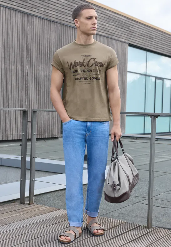Ein Mann steht auf einer Dachterrasse, trägt T-Shirt, Jeans, Sandalen und hält eine Tasche.