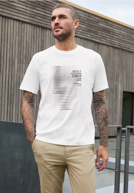 Ein tätowierter Mann mit weißem T-Shirt und beigen Hosen steht draußen vor einer Holzwand