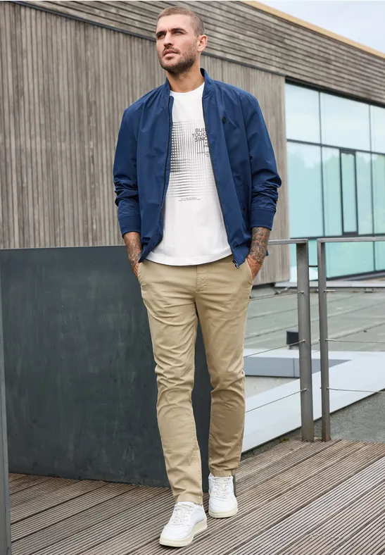 Ein Mann in beige Chinos, weißem T-Shirt und blauer Bomberjacke geht auf einer Holzterrasse.