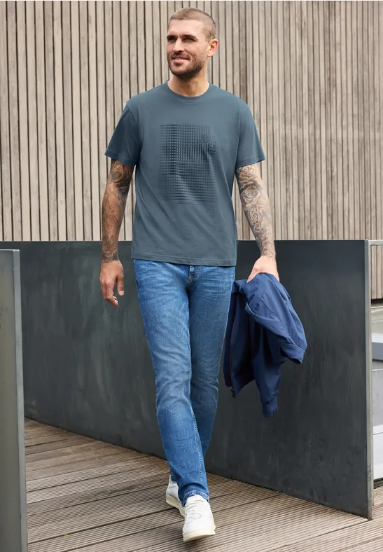 Ein lächelnder Mann in Jeans und T-Shirt hält eine Jacke und geht auf einem Holzsteg vor einer Holzfassade.
