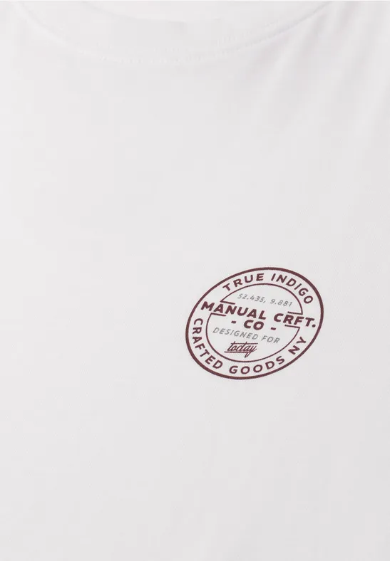 Ein weißes T-Shirt mit ovalem roten Logo-Aufdruck "Manual Crafted Co Limited Goods" auf Brusthöhe.