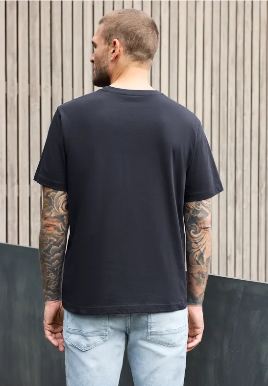 Ein Mann mit Tattoos trägt ein schwarzes T-Shirt und Jeans, steht vor einer Holzlattenwand, Rückenansicht.