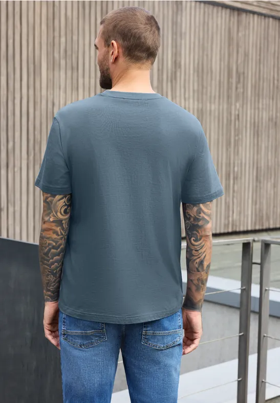 Ein tätowierter Mann von hinten in blauem T-Shirt und Jeans lehnt an einem Holzzaun.