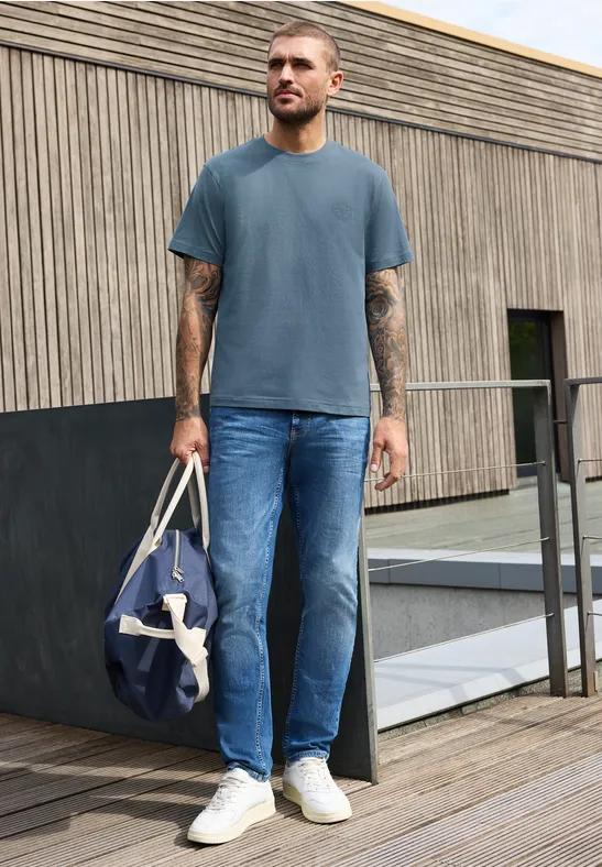 Ein Mann auf einem Holzdeck trägt blaues T-Shirt, Jeans, weiße Sneaker und hält eine blaue Reisetasche.