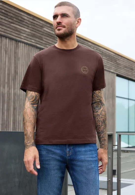 Junger Mann mit braunem T-Shirt und Jeans, tätowierte Arme, steht vor modernem Gebäude.