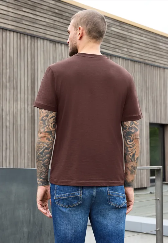 Ein Mann steht mit dem Rücken zur Kamera, trägt braunes T-Shirt und Jeans vor modernem Holzgebäude.