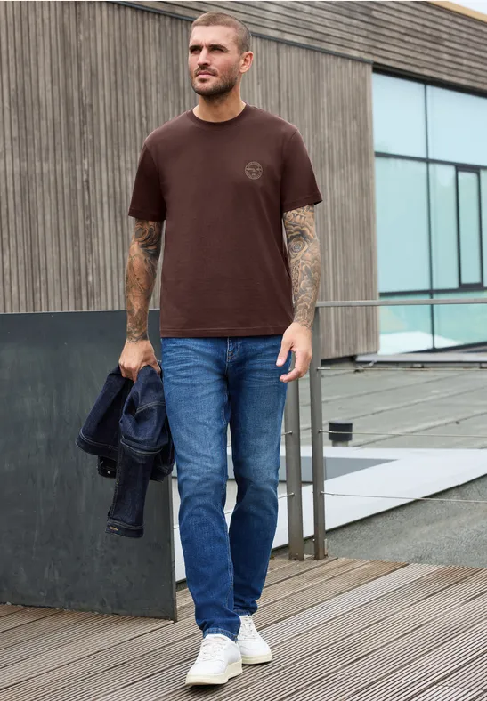 Mann trägt braunes T-Shirt, blaue Jeans und weiße Sneaker, hält Jacke, läuft auf Holzsteg.