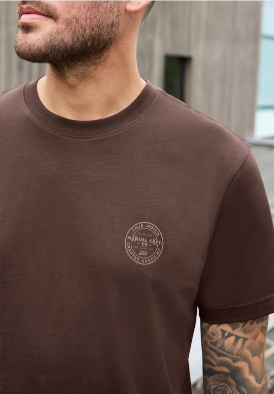 Mann trägt braunes T-Shirt mit kleinem Rundlogo auf der Brust, sichtbarer tätowierter Arm.
