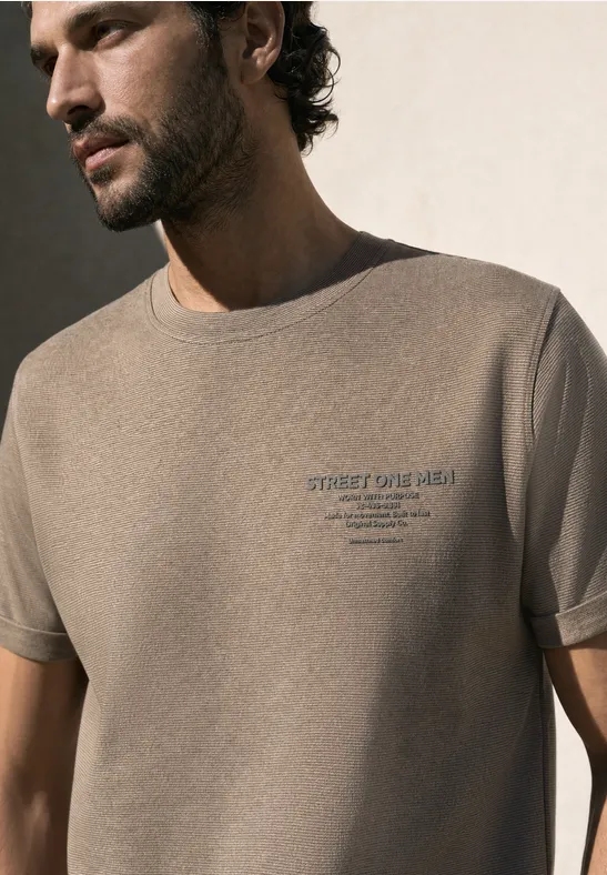 Ein Mann im Profil trägt ein beiges T-Shirt mit dezentem Schriftzug auf der Brust.