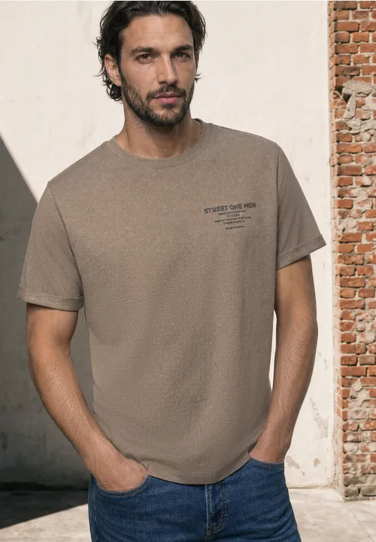 Mann mit beige T-Shirt und blauen Jeans steht entspannt vor einer Ziegelwand.
