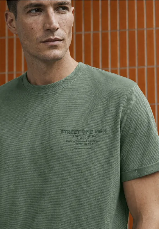 Mann trägt grünes T-Shirt mit kleinem Logo, steht vor orangefarbener Fliesenwand.