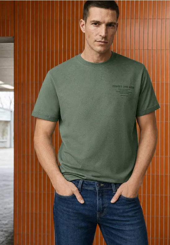 Ein Mann in einem grünen T-Shirt und Jeans steht vor einer orange-braunen Wand mit senkrechten Paneelen.