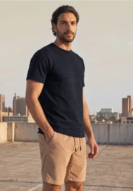 Een man in zwart t-shirt en beige korte broek staat op een dak met stadsgezicht en kijkt in de camera.